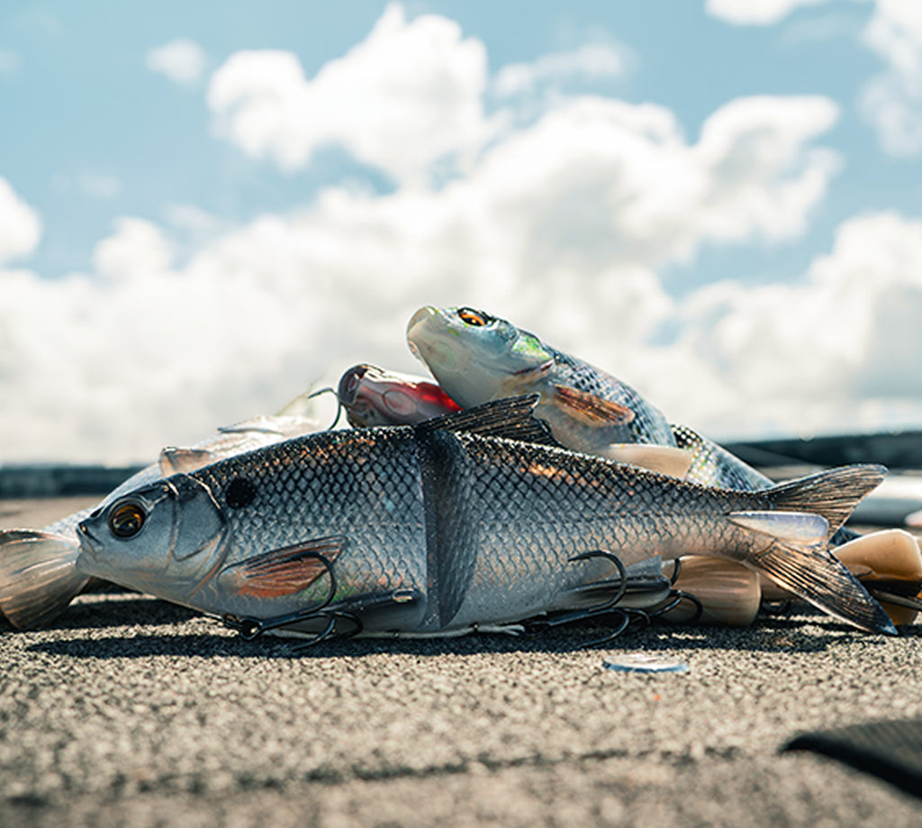 Savage Gear New Swimbaits piled on top of each other on a sandy ground. 